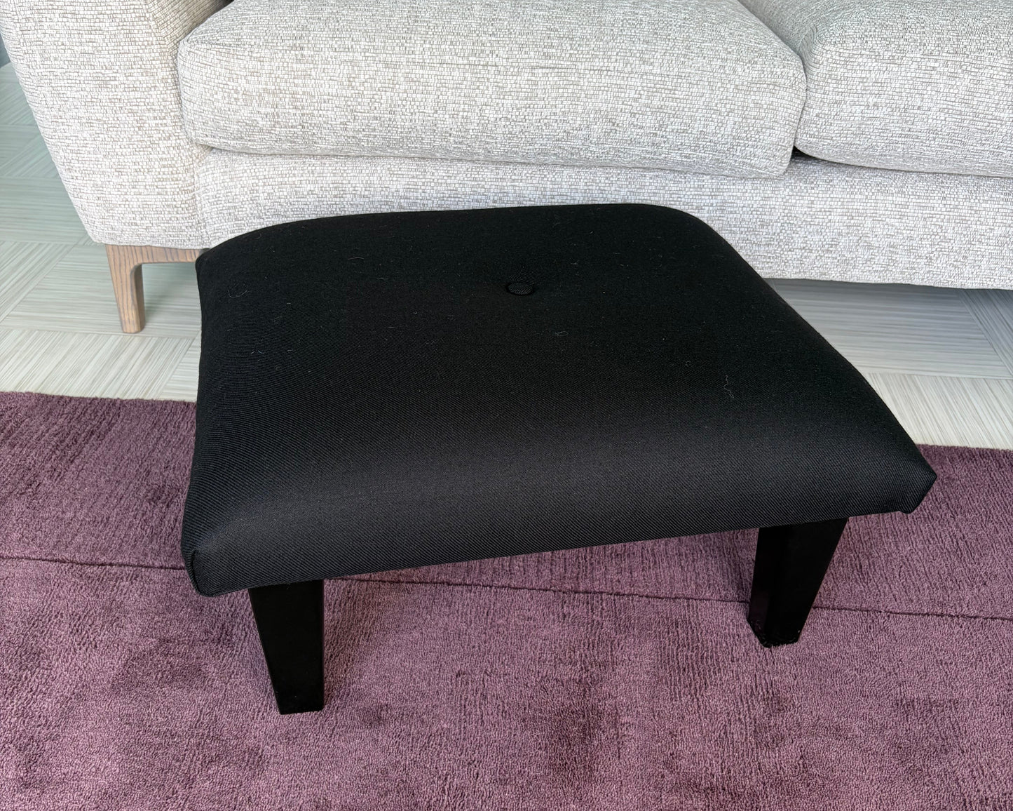 A black upholstered ottoman stool with short legs, shown in a room setting next to a light gray sofa and a purple textured rug.