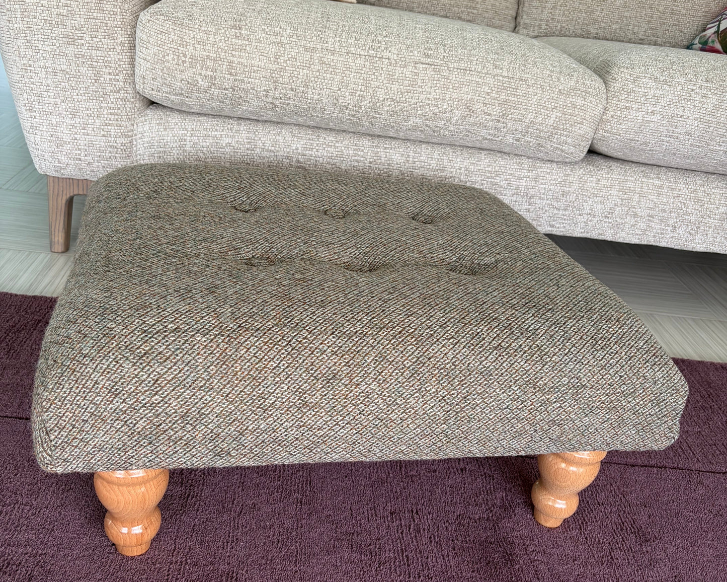 A square tufted ottoman stool upholstered in a textured gray herringbone fabric with turned wooden legs, placed on a dark purple rug in front of a light gray sofa.