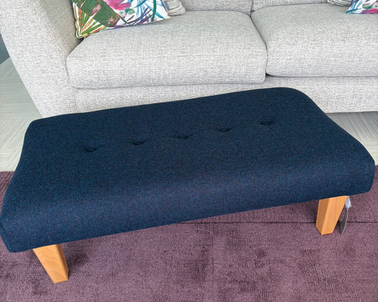 A dark navy blue upholstered ottoman with light wood legs, positioned in front of a light gray fabric sofa on a purple rug.