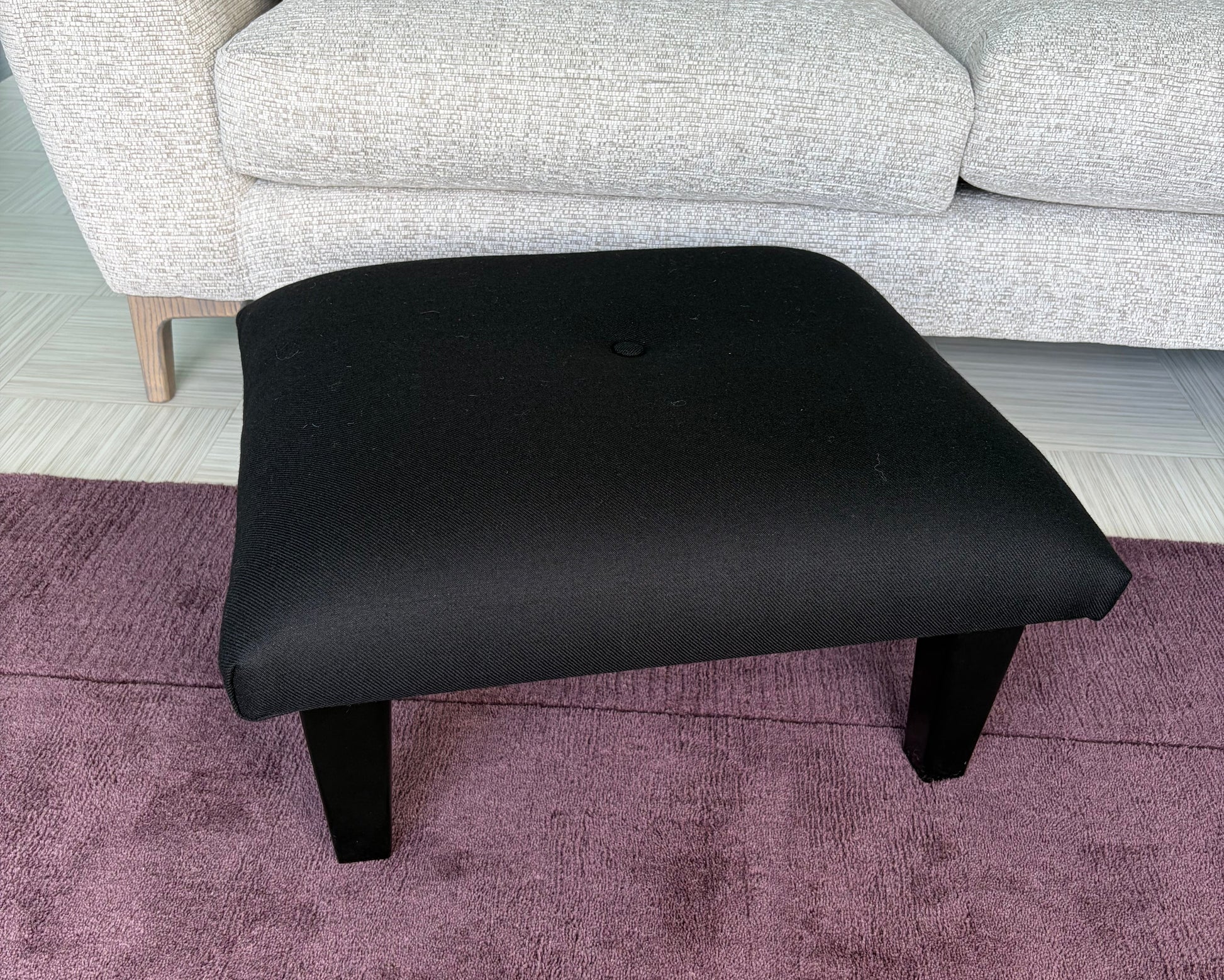 A black upholstered ottoman stool with short legs, shown in a room setting next to a light gray sofa and a purple textured rug.
