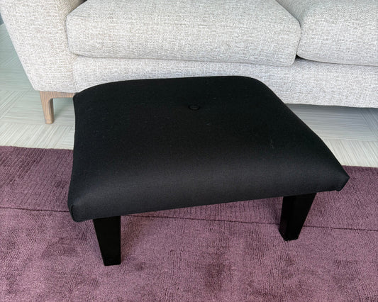 A black upholstered ottoman stool with short legs, shown in a room setting next to a light gray sofa and a purple textured rug.