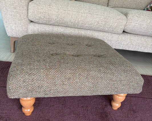A square tufted ottoman stool upholstered in a textured gray herringbone fabric with turned wooden legs, placed on a dark purple rug in front of a light gray sofa.